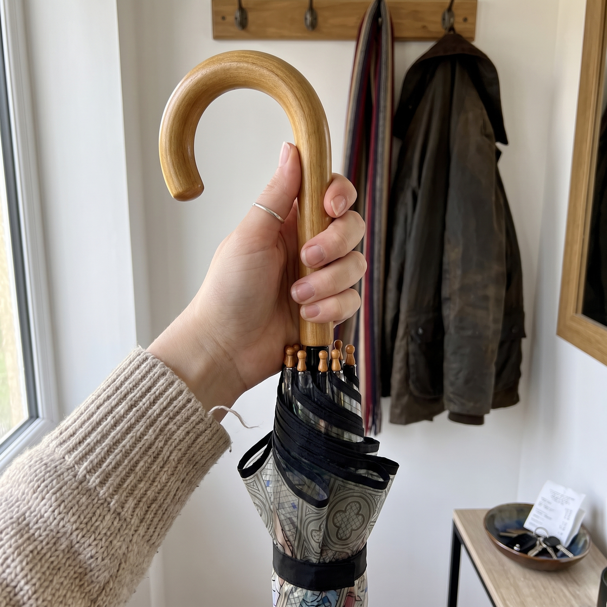 A hand holding a closed umbrella with a curved light wood handle and a patterned fabric body in an indoor setting with a coat rack and jacket in the background.